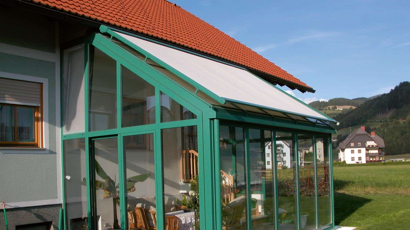 Glaswintergarten mit grünem Rahmen an einem Haus mit schrägem weißem Dach vor dem Hintergrund einer ruhigen Landschaft.